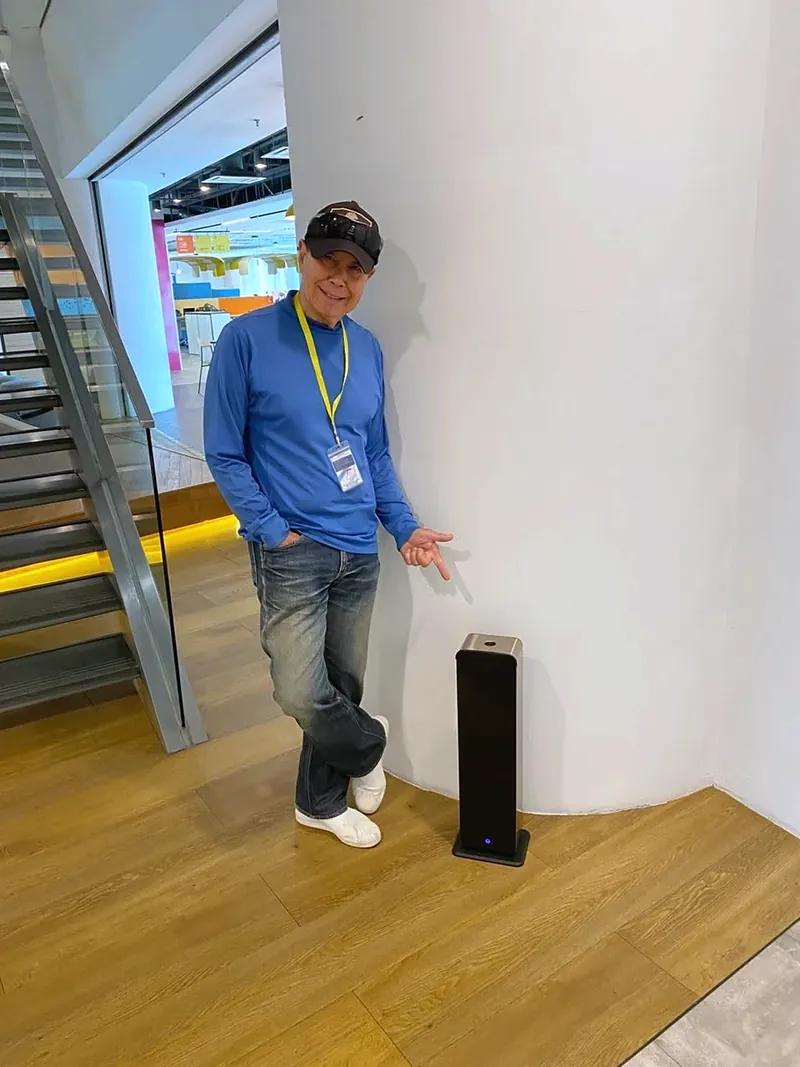 John Lee, Scent Up's partner in Malaysia, pointing to a black tower scent diffuser placed against a wall in a modern office interior.